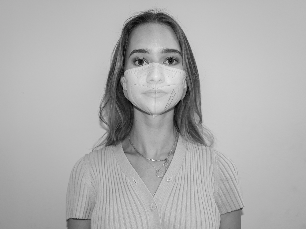 Auf dem Bild ist eine junge Frau zu sehen, die eine Mund-Nasen-Schutz trägt. Der Mund-Nasen-Schutz ist jedoch durchscheinend, sodass das Gesicht der Frau darunter noch zu sehen ist.