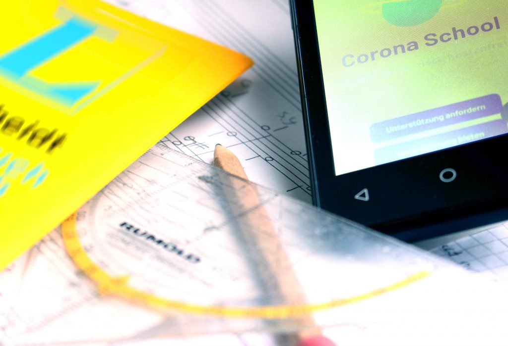Ein Wörterbuch, ein Geodreieck, ein Bleistift und ein Smartphone, das den Schriftzug "Corona School" anzeigt, liegen auf einem Blatt Notenpapier.