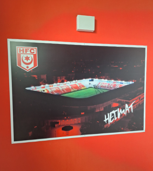 Das Foto zeigt ein eingerahmtes Bild des HFC-Stadions auf einer rot gestrichene Wand. Der Bilderrahmen ist weiß gefärbt. Der Inhalt des Bildes ist das bei Nacht leuchtende Stadion des Fußballclubs. Die darum liegende Fläche erstrahlt in rotem Licht. Rechts unten im eingerahmten Bild befindet sich der Schriftzug „Heimat“. In der oberen linken Ecke ist das Logo des halleschen Fußballvereins abgebildet. 