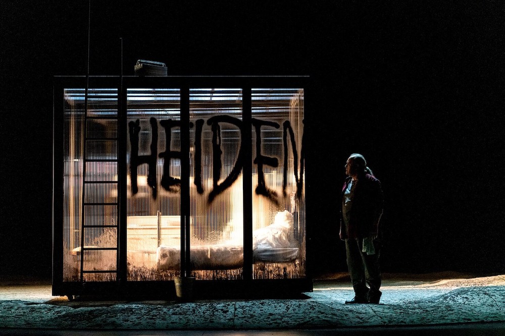 Das Bild ist eine Fotografie aus einer Szene von "Manru". Es ist sehr dunkel und eine Art beleuchteter Container mit farblosen Wänden ist zu sehen. Es ist die Behausung von Manru und Ulana. Daneben steht Manru, es ist nur seine Silhouette zu erkennen. Die Wände der Behausung wurden beschmiert.