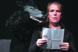 Auf dem Bild ist eine Person mit Zettel in der Hand. Sie schaut ausdruckslos, vielleicht ein wenig erschrocken gerade aus. Neben ihr ist die Schnauze der Handpuppe eines schwarzen Hundes zu sehen. Bedrohlich nahe ist die Hundenase ihrem Gesicht.